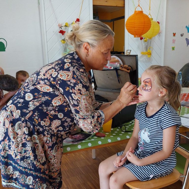 Kinderschminken. Eine Frau schminkt ein Kind. Dieses bekommt einen Schmetterling um die Augen gezeichnet.